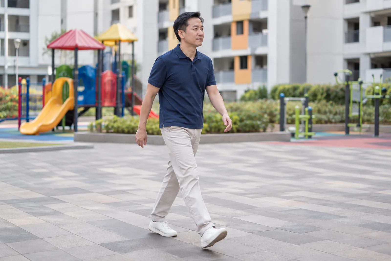 A man walks briskly across an open housing-estate plaza with a playground and fitness equipment behind him.