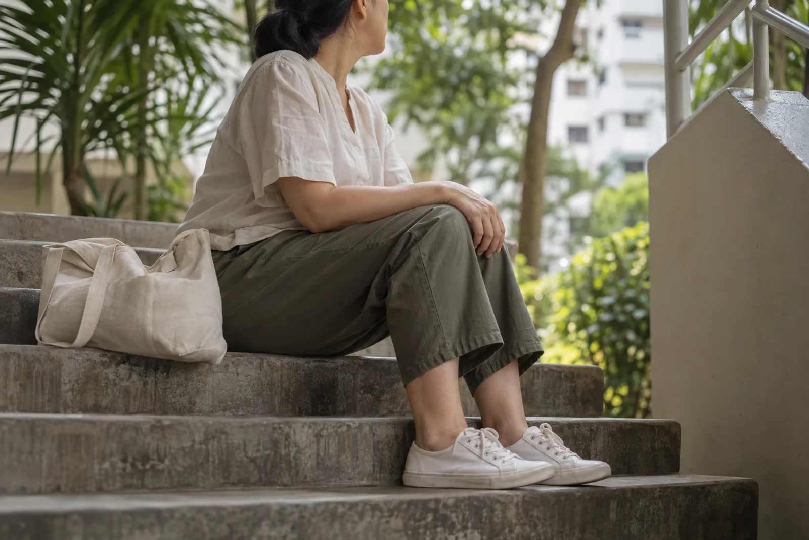 An adult sits thoughtfully on an outdoor residential staircase with one hand resting on the knee and a canvas bag beside them.