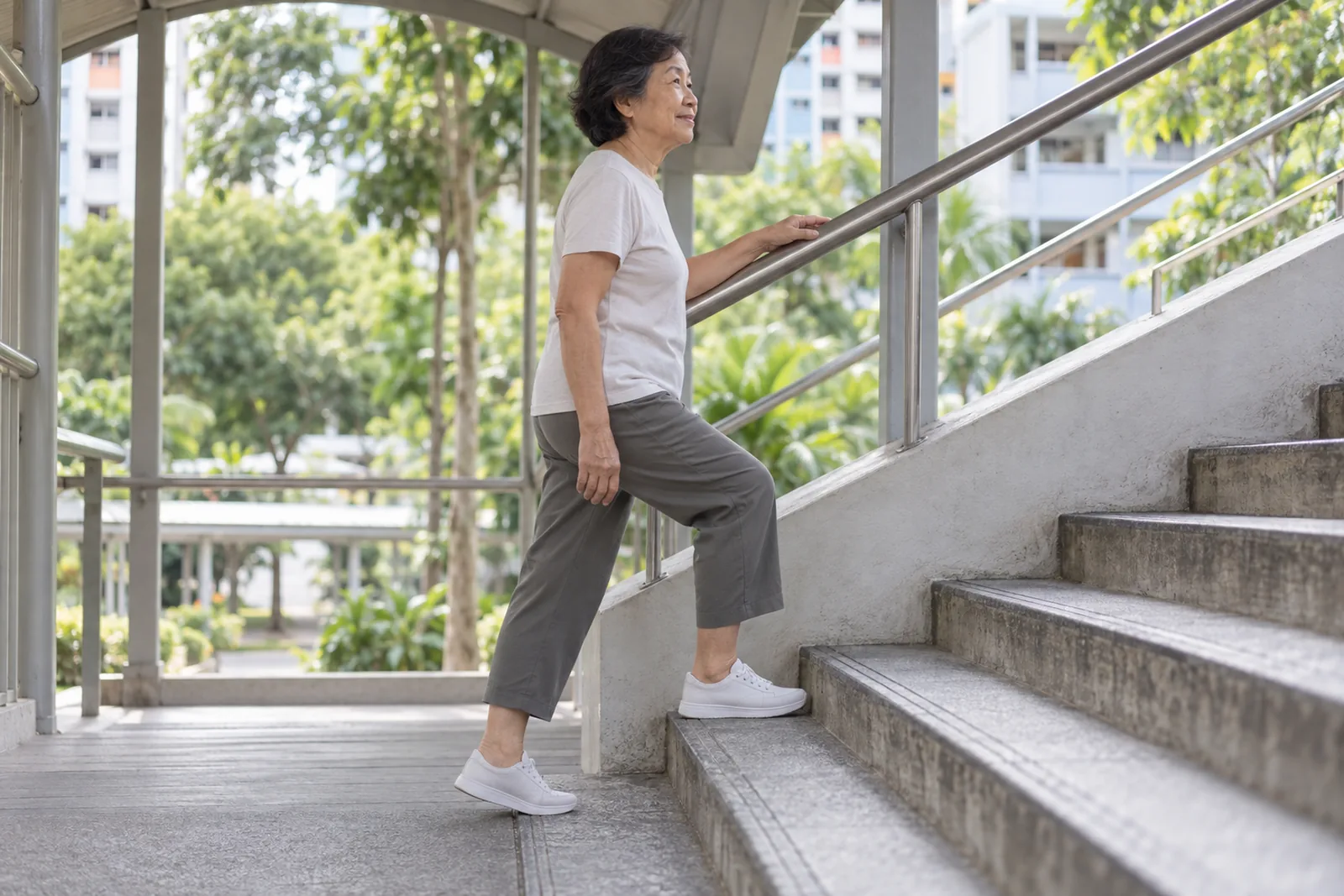An older woman climbs an HDB overhead bridge staircase with one hand resting lightly on the rail.