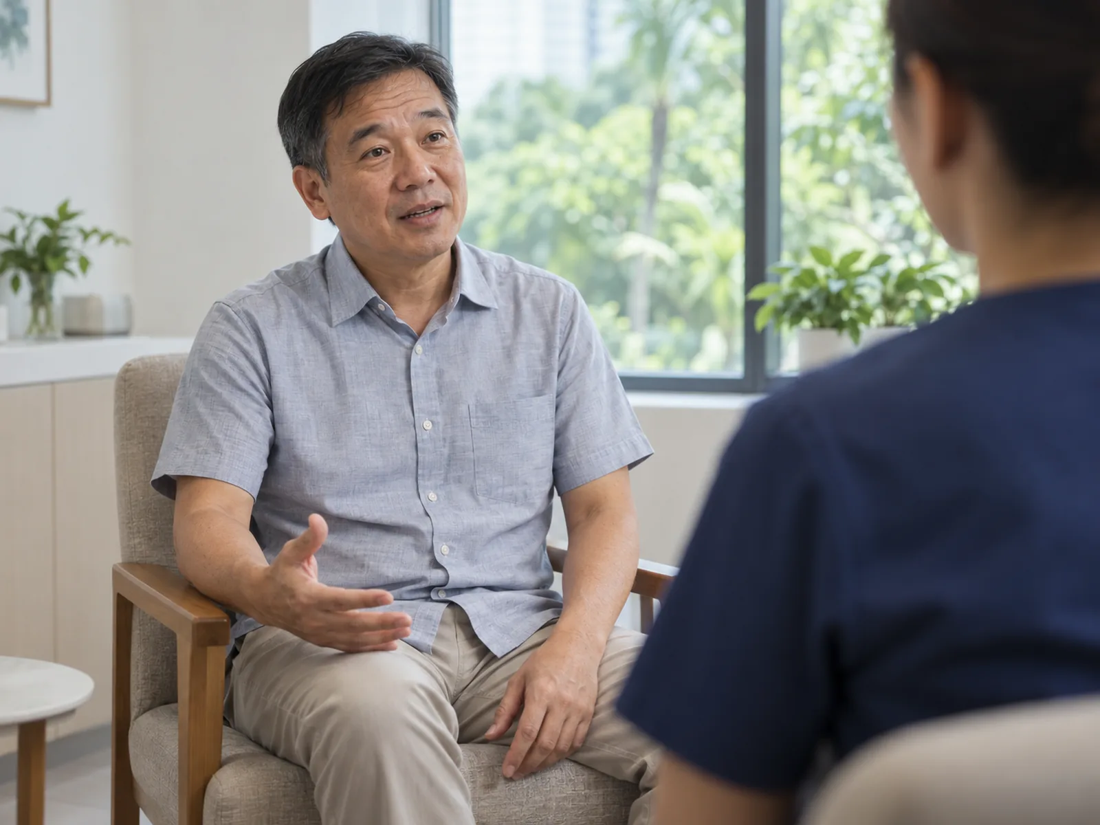 A seated man gestures while speaking with a clinician in a bright consultation room overlooking greenery.