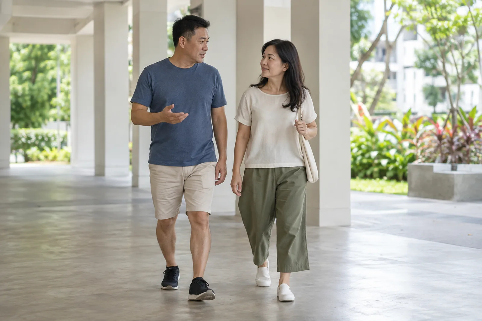 A couple walk through a bright housing-estate void deck while talking calmly with each other.