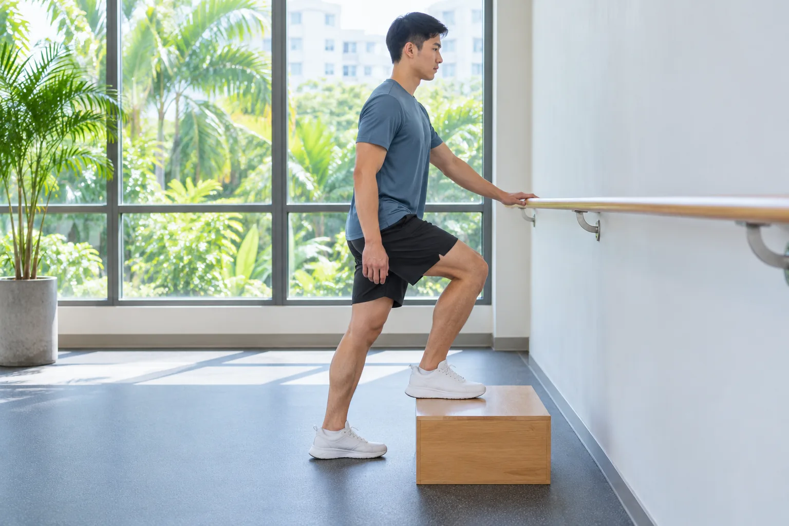 A young man performs a controlled step-up on a low wooden exercise box beside a wall rail in a bright community gym.