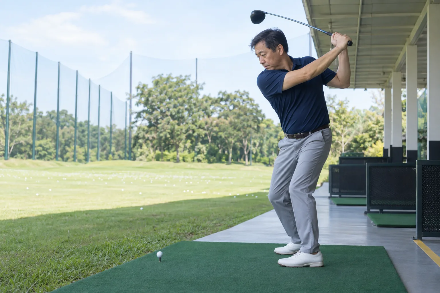 A man stands at the top of his golf backswing in a driving-range bay.