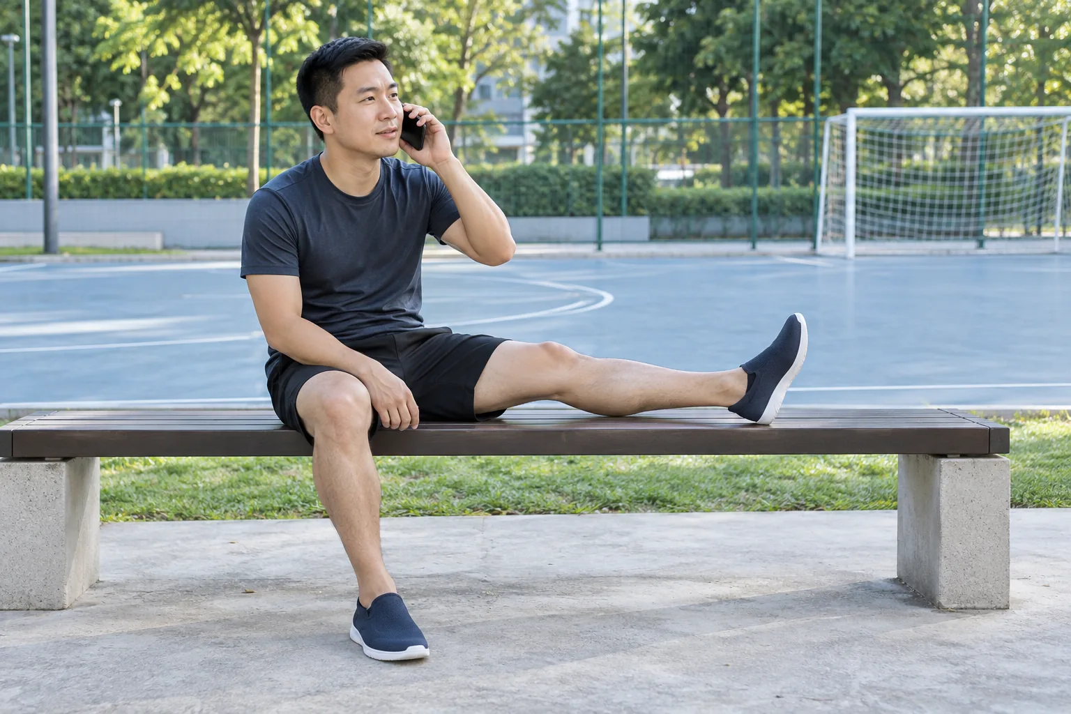 A man sits beside a futsal court with one leg stretched along a bench and speaks calmly on a telephone.