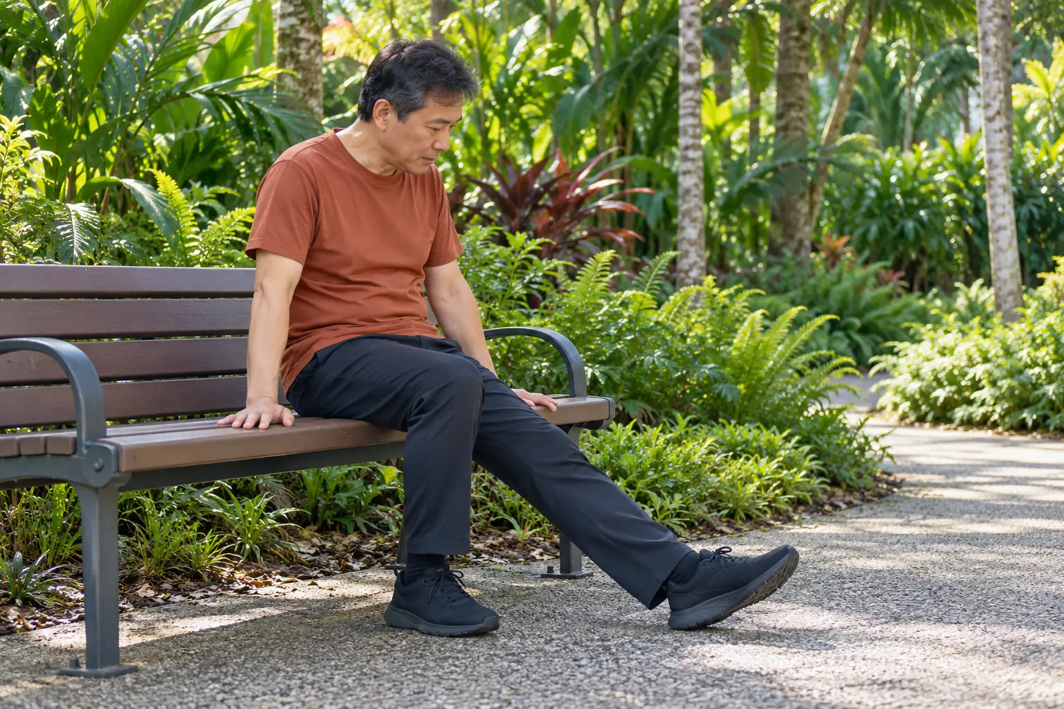 A man sits on a park bench and looks down at his outstretched leg with his hands resting on the bench.