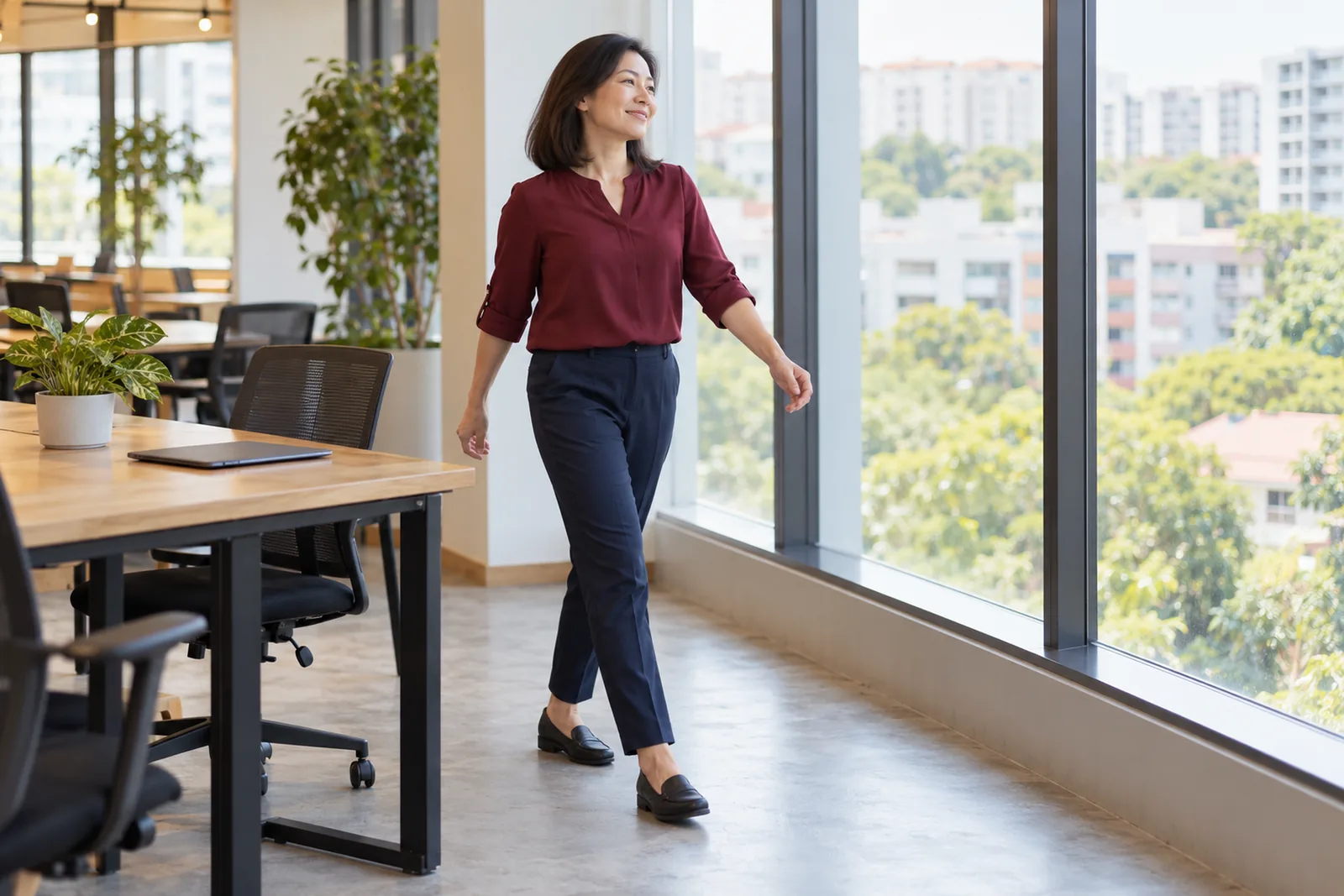 A woman walks from her desk towards a bright window overlooking ordinary housing blocks and tree tops.