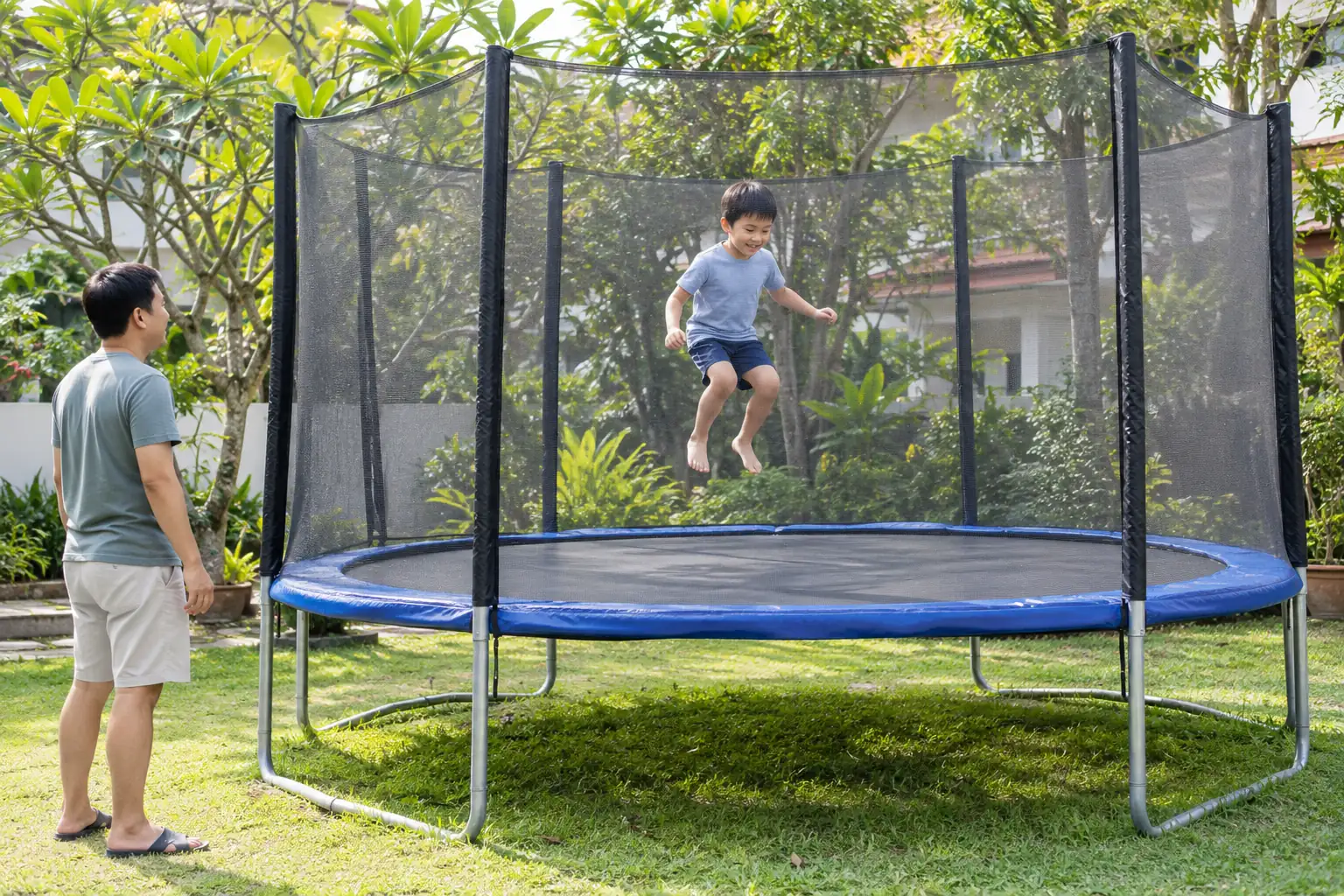 A boy bounces inside a net-enclosed trampoline while a parent watches beside it.